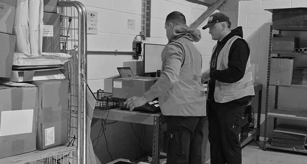Two workers in a warehouse setting, one using a computer and the other observing.