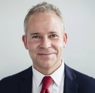 Man wearing a dark suit with a red tie against a plain background
